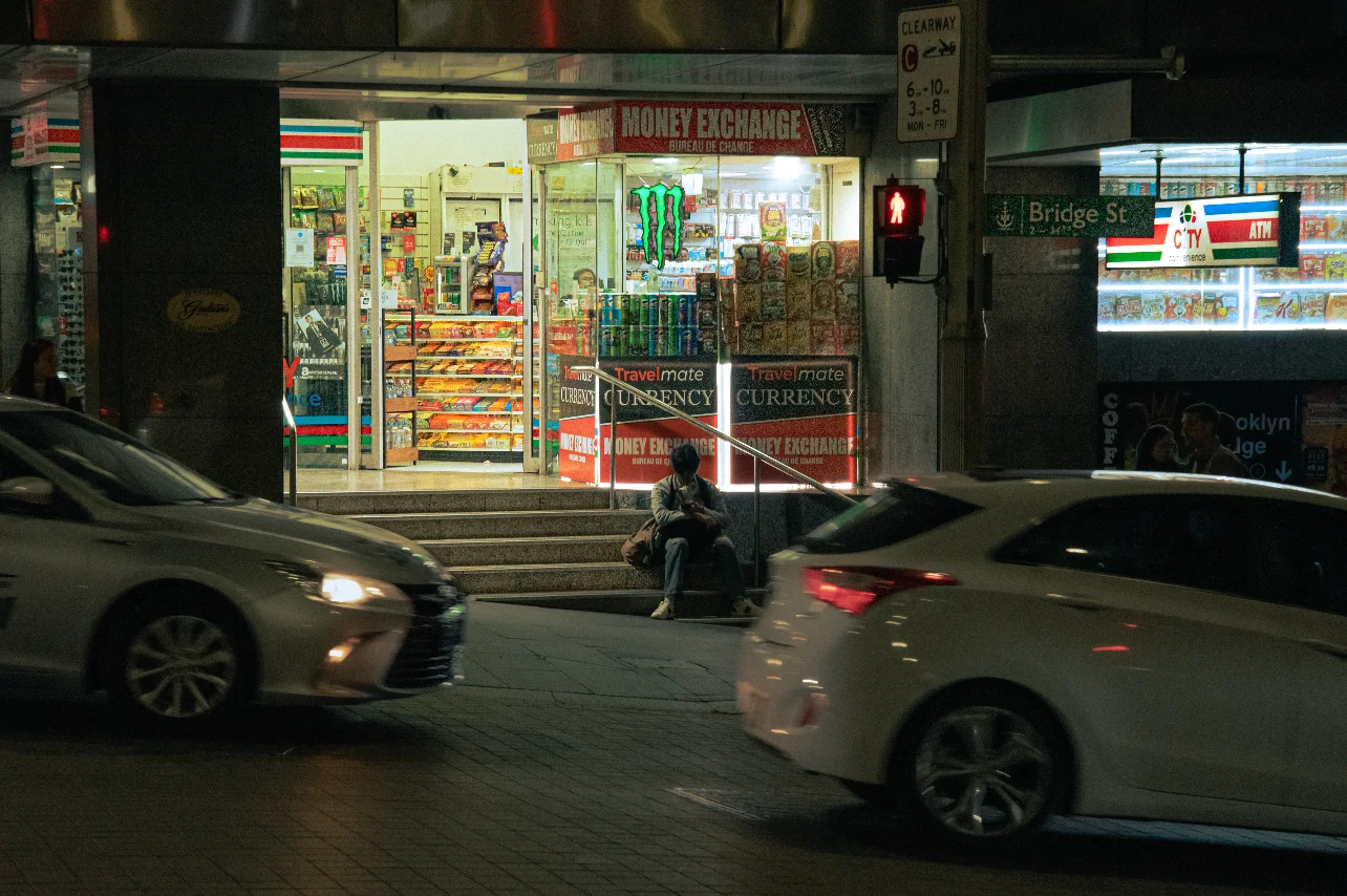 Edge of frame, with a maximum depth of field, with cars entering and leaving the frame with a man sitting in front of a convinience store in the background